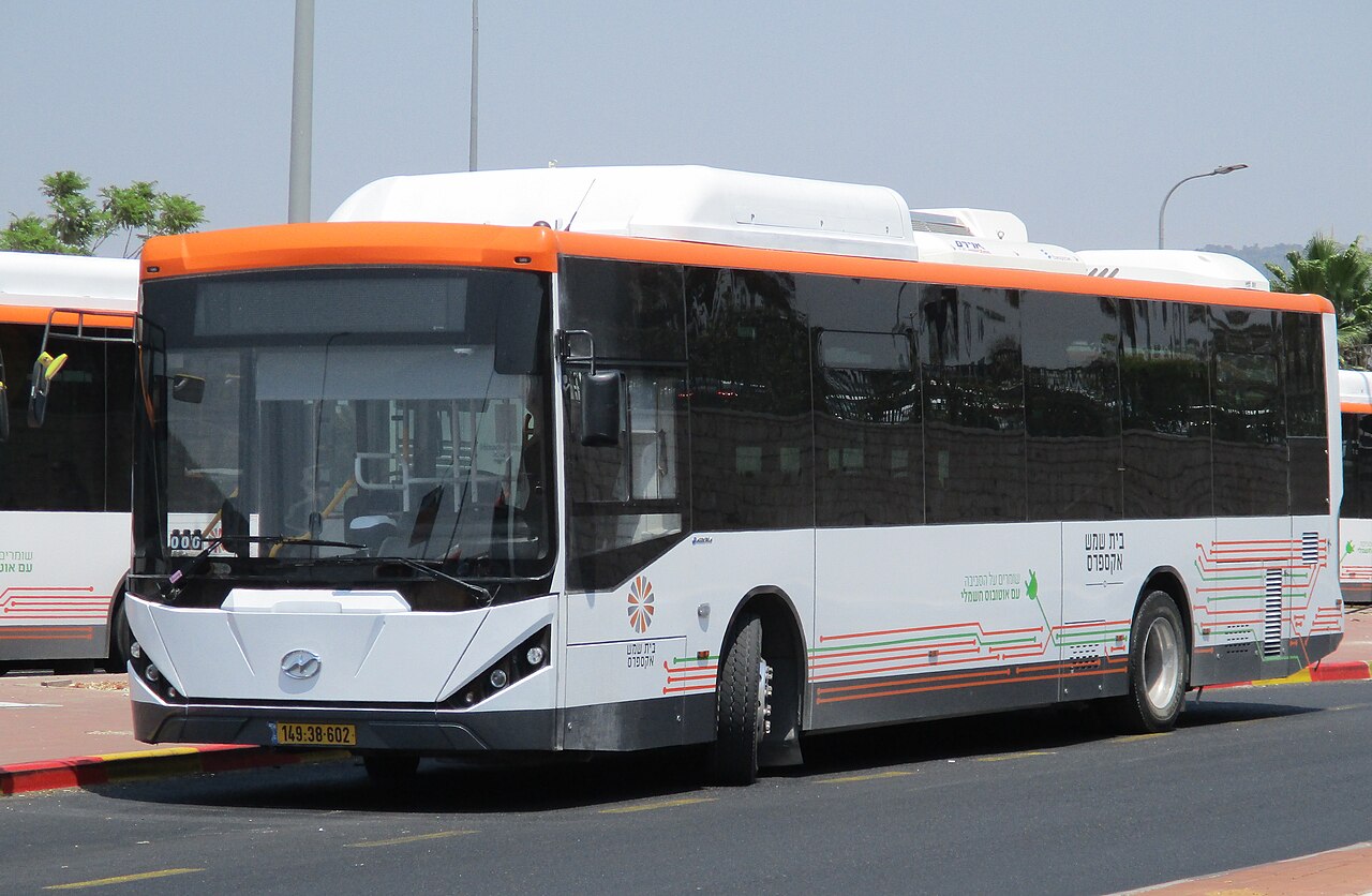 אוטובוס נוסעים לבן עם פס אדום בחלקו העליון, נוסע על רקע שמים אפורים.
