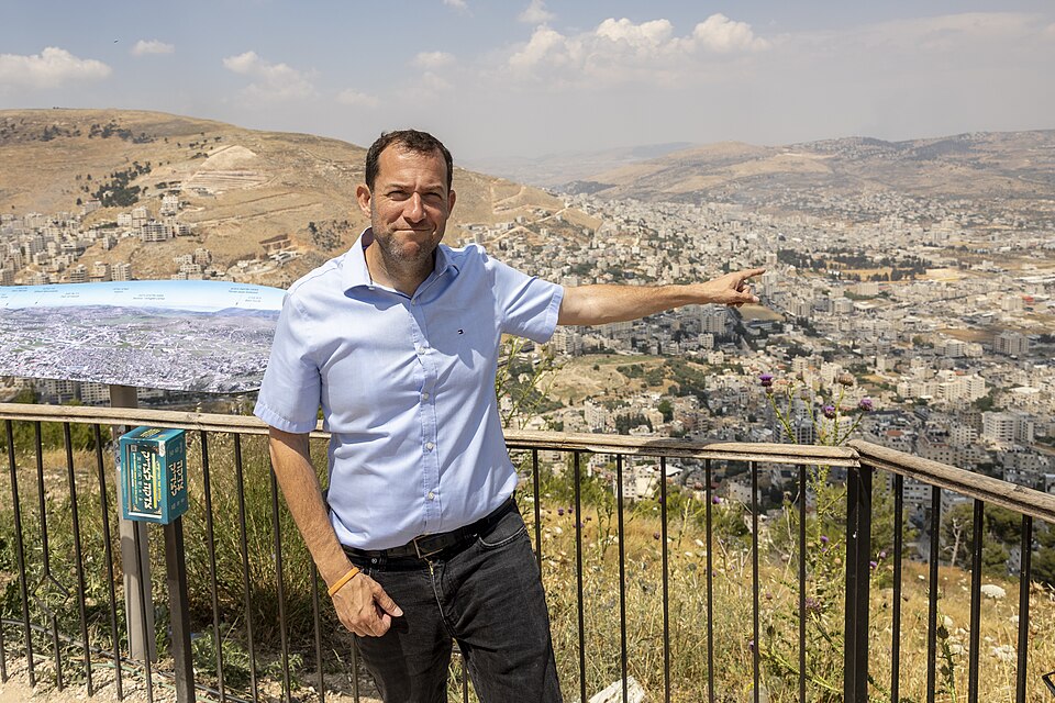 איש חובש חולצה בצבע תכלת כהה, מרים את ידו הימנית, מול תפיסת נוף עירונית המשקיפה על גבעות.