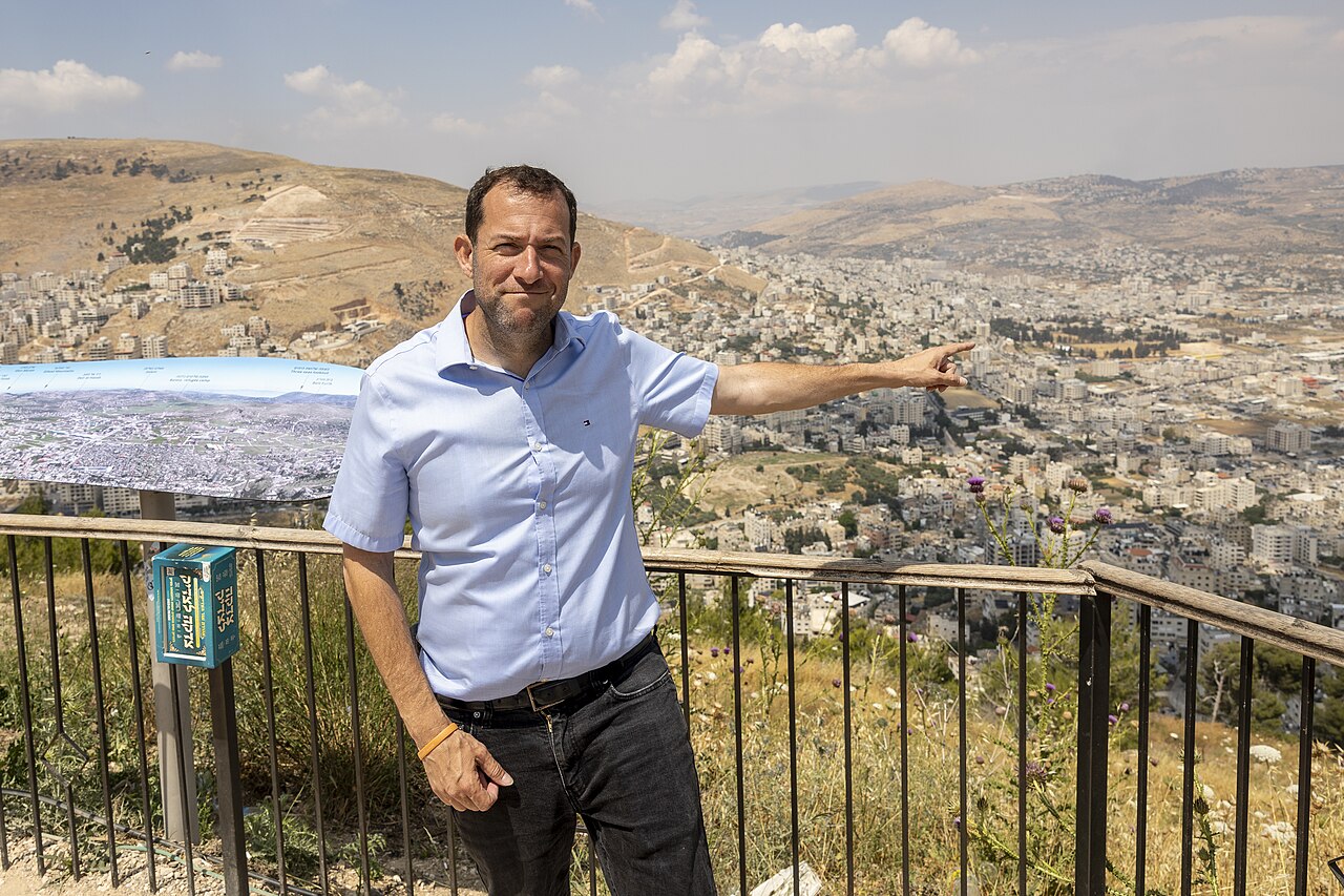 איש חובש חולצה בצבע תכלת כהה, מרים את ידו הימנית, מול תפיסת נוף עירונית המשקיפה על גבעות.