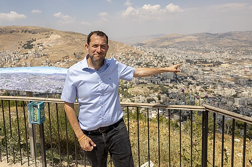 איש חובש חולצה בצבע תכלת כהה, מרים את ידו הימנית, מול תפיסת נוף עירונית המשקיפה על גבעות.