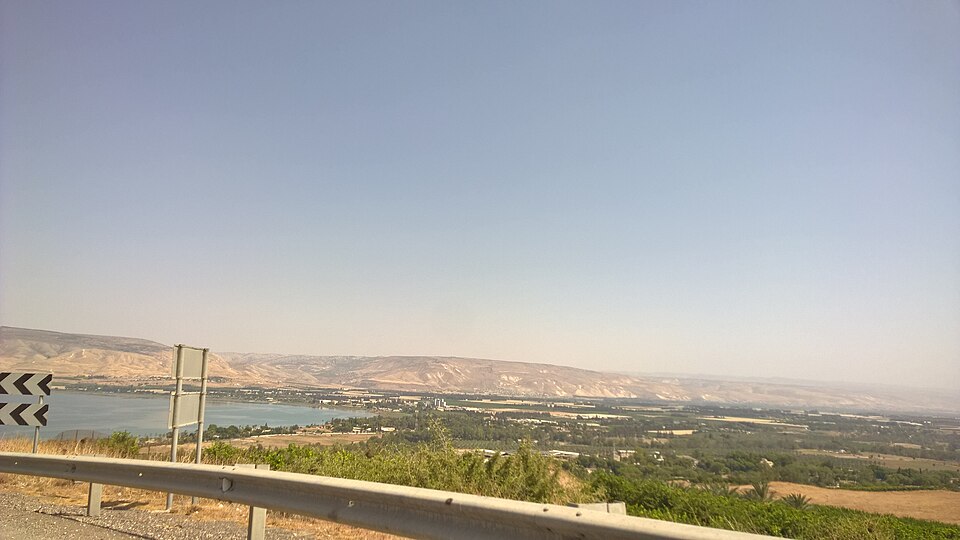 נוף פנורמי של עמק, כנראה עמק הירדן, עם גוף מים, שדות ורכס הרים ברקע, לצד כביש עם גדר בטיחות.