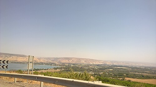 נוף פנורמי של עמק, כנראה עמק הירדן, עם גוף מים, שדות ורכס הרים ברקע, לצד כביש עם גדר בטיחות.