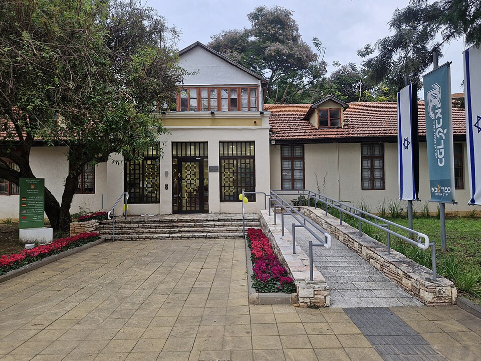 בניין ציבורי עם דלתות כניסה, מדרגות, משטח גישה עם שיפוע קל, דגלי ישראל, עצים, פרחים ומדרכה.