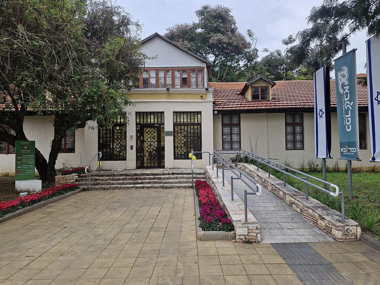 בניין ציבורי עם דלתות כניסה, מדרגות, משטח גישה עם שיפוע קל, דגלי ישראל, עצים, פרחים ומדרכה.