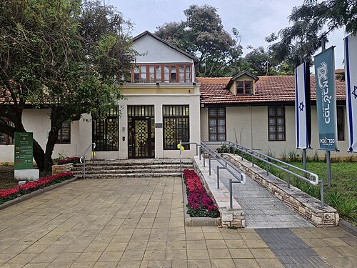 בניין ציבורי עם דלתות כניסה, מדרגות, משטח גישה עם שיפוע קל, דגלי ישראל, עצים, פרחים ומדרכה.