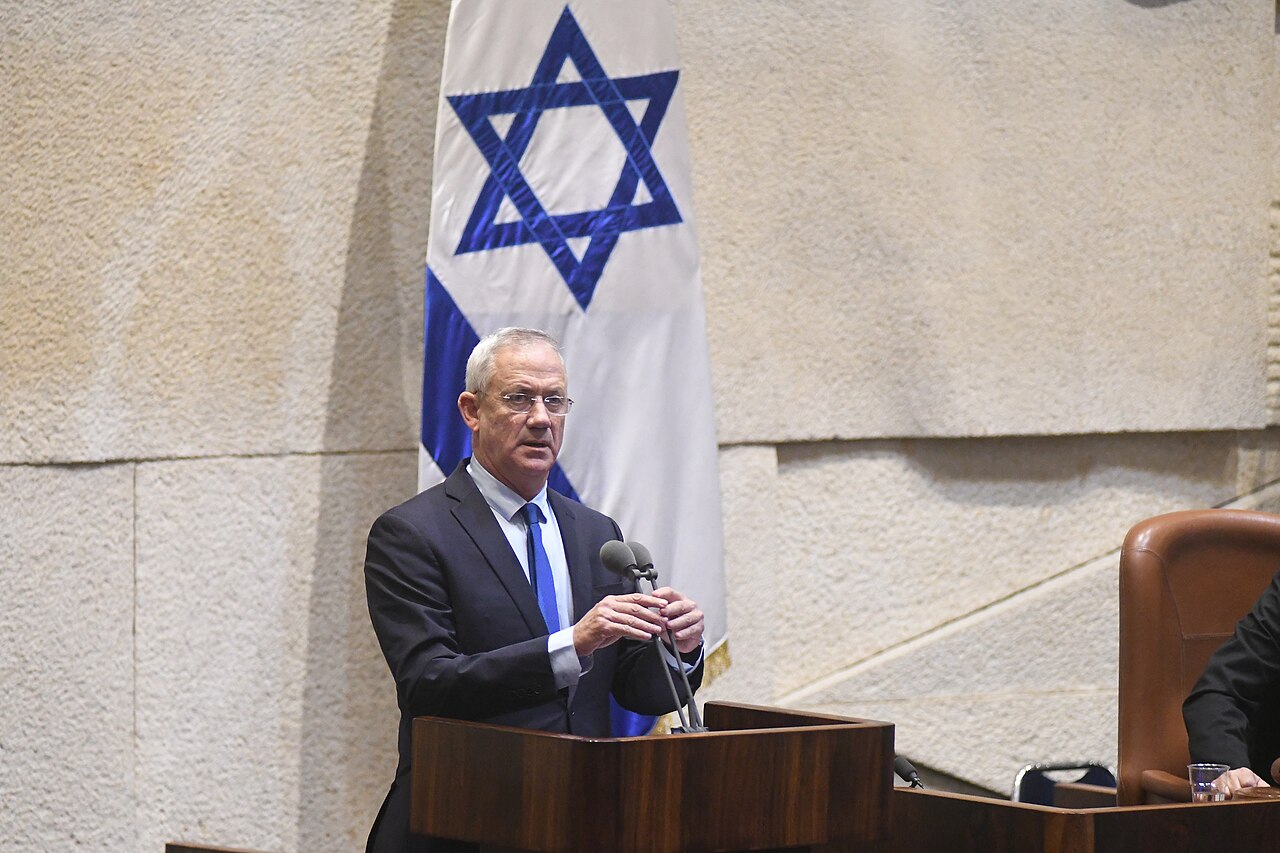 איש מבוגר יותר, בחליפה כהה, עומד ומדבר בעזרת מיקרופון, מול דגל ישראל.