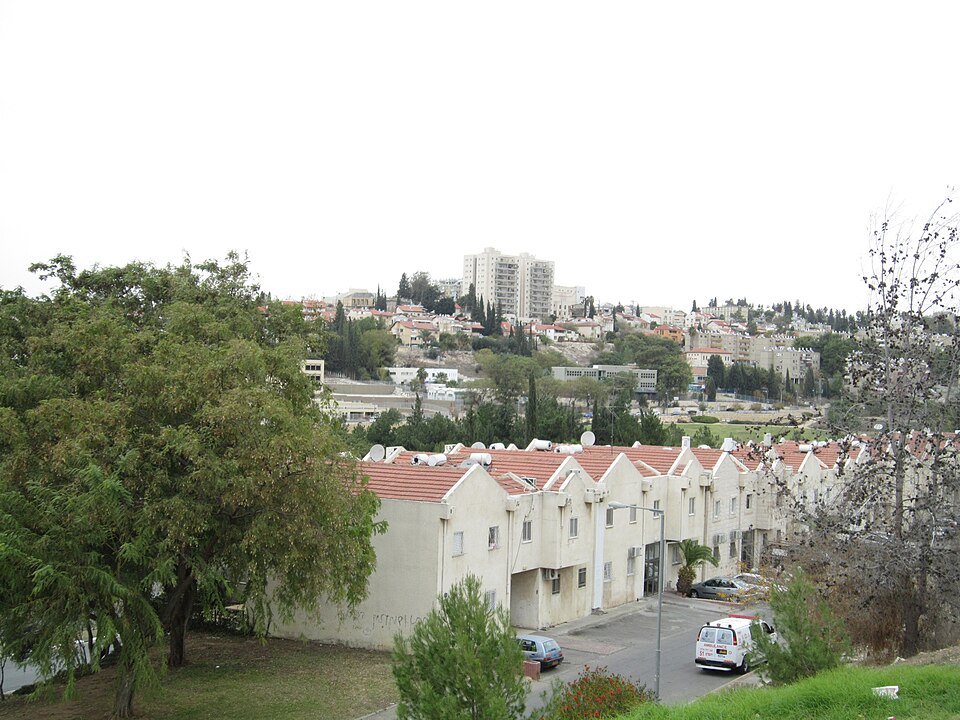 עשרים וחמישה בתים לבנים עם גג אדום, חלקם עם חניה, נמצאים ברקע תמונת נוף עירונית עם עצים.