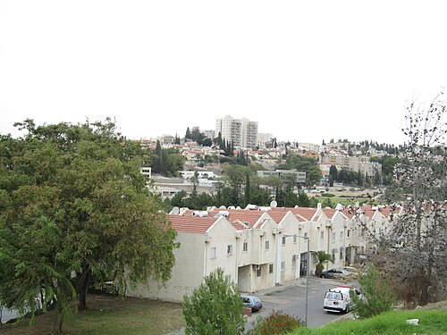עשרים וחמישה בתים לבנים עם גג אדום, חלקם עם חניה, נמצאים ברקע תמונת נוף עירונית עם עצים.