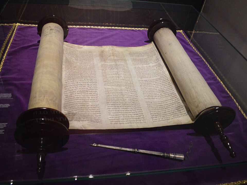 התמונה מראה מגילה עתיקה עם טקסט כתוב עליה, המוצגת בתיבה זכוכית עם רקע סגול.