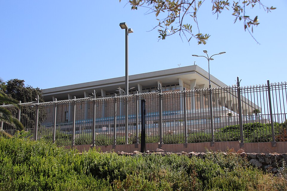 מבנה גדול ומוארך, עם גג שטוח, מוקף גדר ברזל, ברקע שמים כחולים.