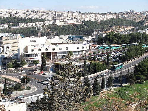 תמונת אוויר של איזור עירוני עם מבנים, עצים, כבישים, חניון ואוטובוסים.