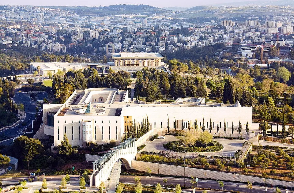 בניין גדול ומלבני עם גג שטוח, גדרות, עצים, שיחים וגשר, מוקף בבניינים, עצים ושדרות.
