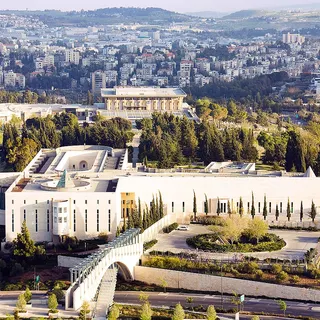 בניין גדול ומלבני עם גג שטוח, גדרות, עצים, שיחים וגשר, מוקף בבניינים, עצים ושדרות.